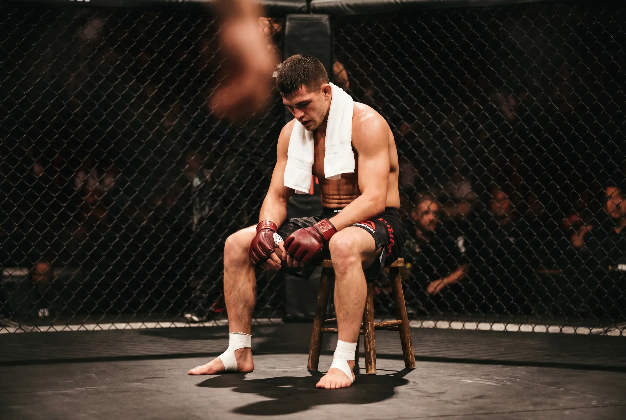 UFC fighter sitting on the corner stool between rounds inside the octagon, head down, white towel around the neck, MMA gloves resting on the knees