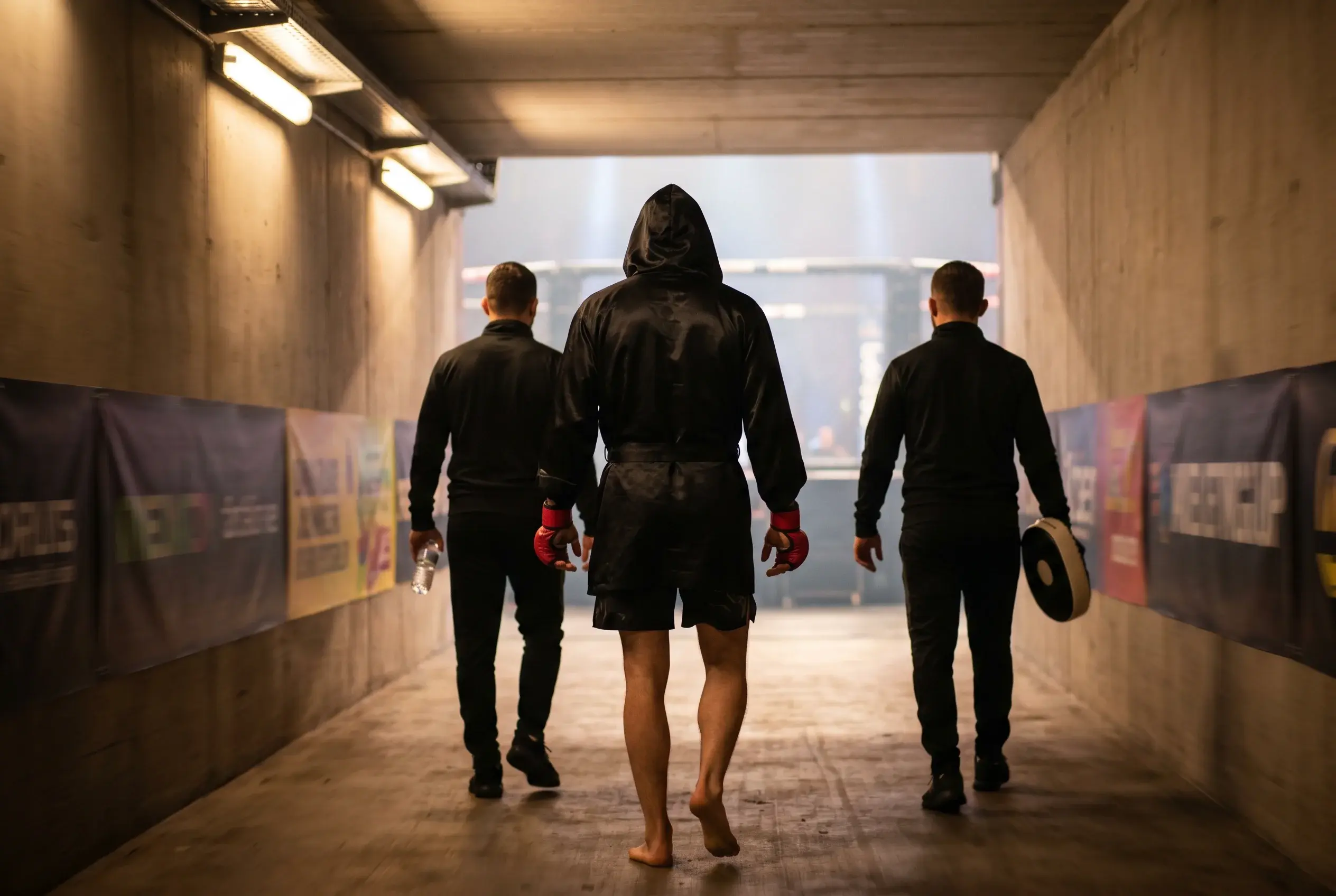 UFC fighter walking down the dim arena tunnel toward the octagon, hooded, with the coach and cornermen following close behind